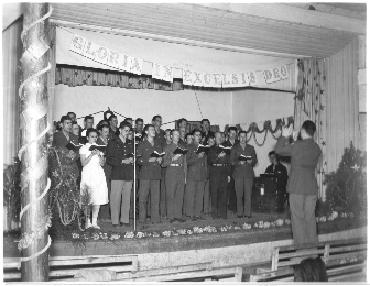 1944 Chrismas Choir on stage, with leader standing on bench in front and accompanyist far right.  Decorated Christmas trees on either side of the stage, and overhead banner reading 'Gloria In Excelsis Deo.