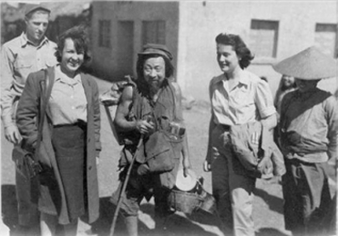Eileen Denney and Betty Ringland stand on each side of a roving food vendor with walking stick in his right hand; all facing camera. He holds buckets of rice and a plate in left hand, had has carry sacks with unidentifiable objects on his back and chest.